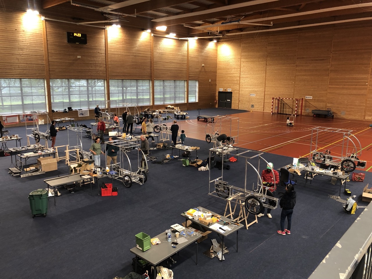 Des personnes montent des Vhélios dans un grand gymnase, entourées de pièces, d’outils et de structures en cours de construction.